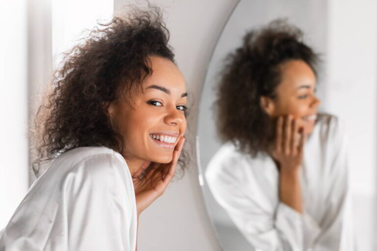 Attractive young African American lady with soft moisturized complexion touches her face during daily skincare routine in bathroom, posing near mirror in silk bathrobe. Selective focus